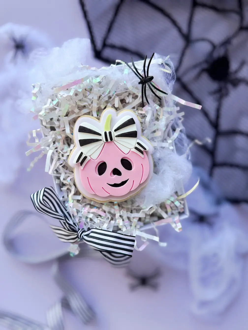 Decorated Halloween cookie shaped like a pink pumpkin with a smiling face and a large black-and-white striped bow.