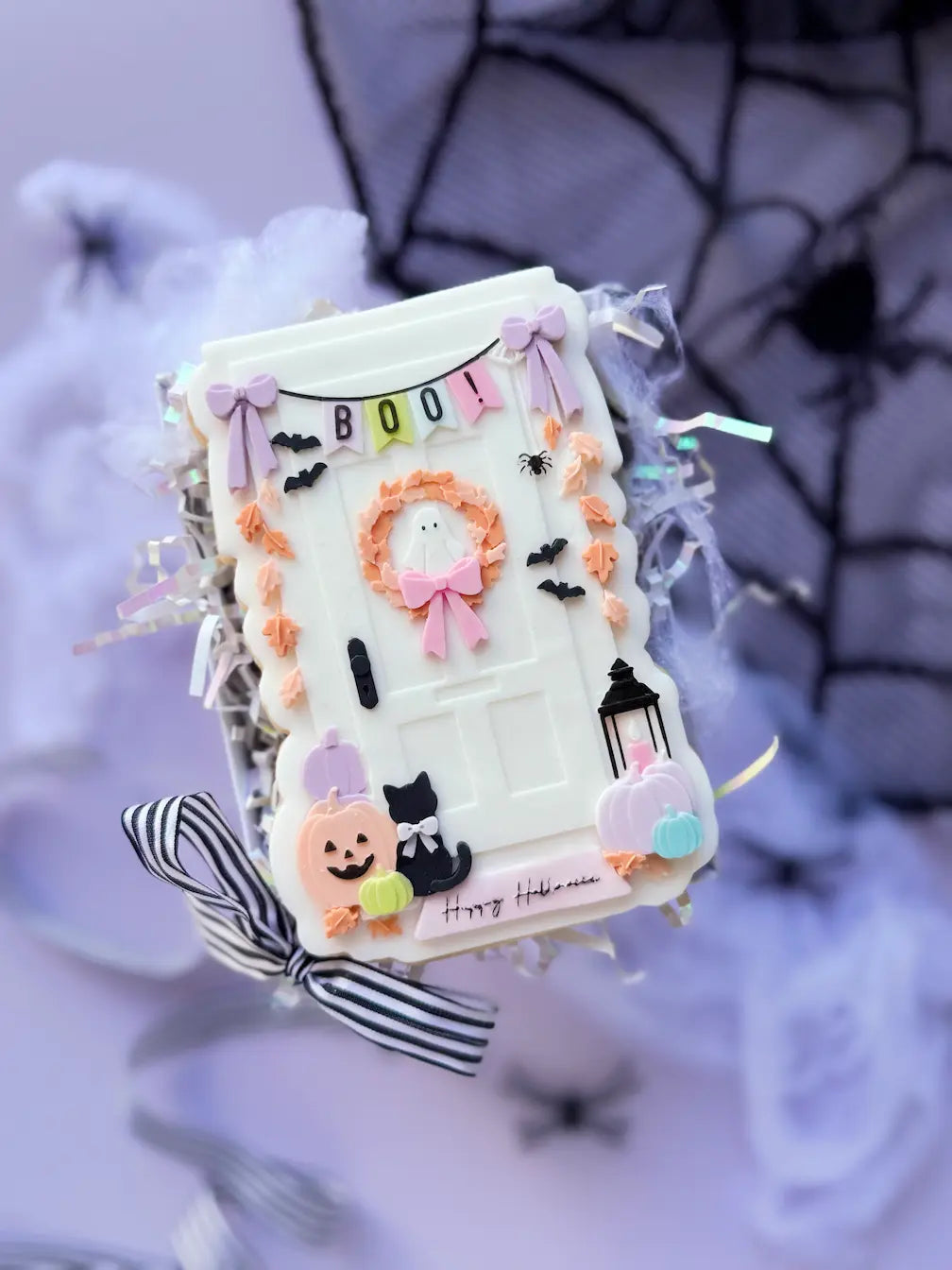 Decorated cookie shaped like a white Halloween door with a BOO banner, ghost wreath, pumpkins and a black cat.