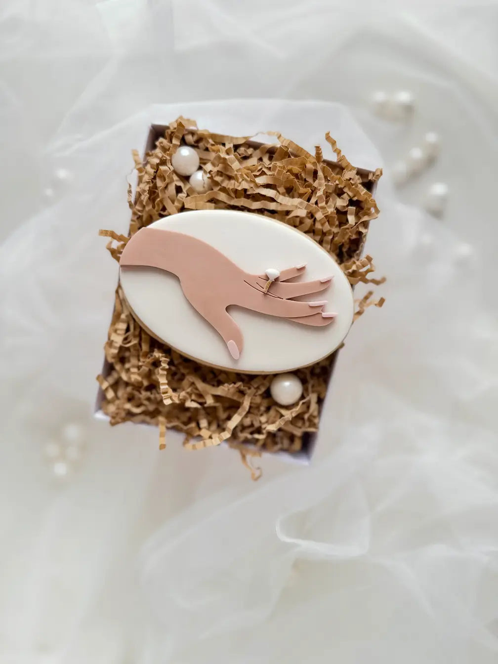 Ovaler weißer Keks mit roséfarbener Hand, hellen Fingernägeln und Verlobungsring.