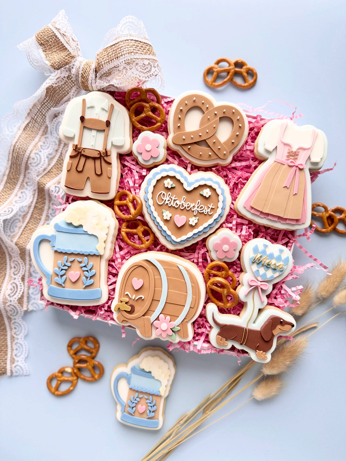 Oktoberfest-Kekssortiment mit Dirndl, Lederhose, Brezel, Bierkrug, Bierfass, Herz und Dackel auf pinkem Füllpapier.
