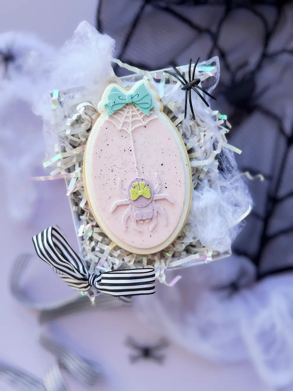 Oval pale pink fondant cookie with a spider, a spiderweb and a turquoise bow.