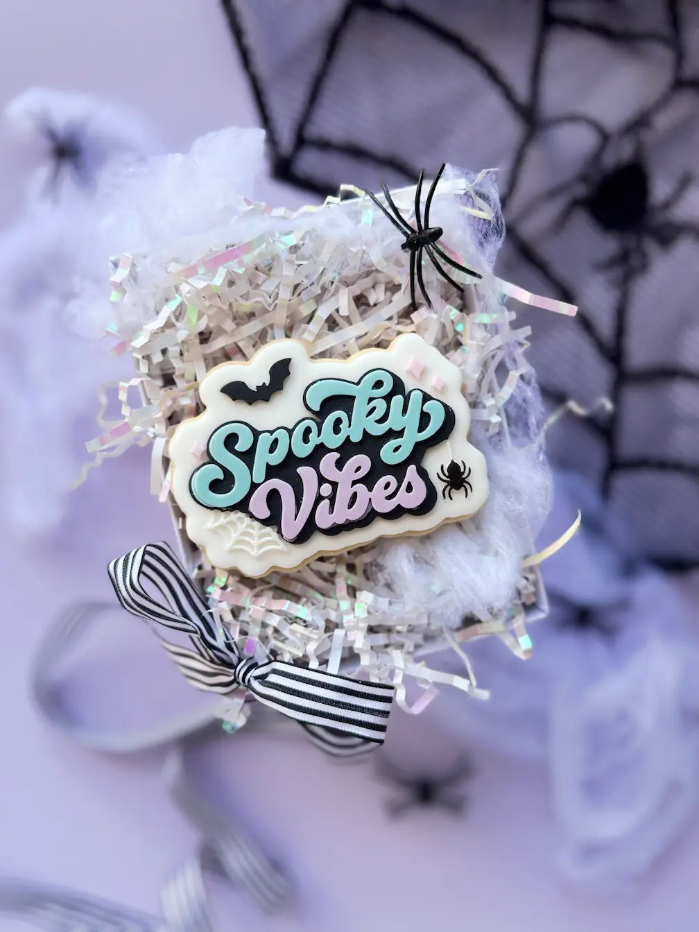 Decorated cookie with turquoise and lilac Spooky Vibes lettering, a black bat, a spider and a white spiderweb.